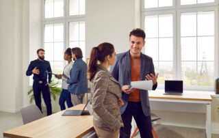 Colleagues collaborating in a modern office space with large windows, discussing documents and ideas. In the foreground, two professionals engage in a conversation, while another group of three stands in the background having a discussion. The setting is bright and professional, fostering teamwork and communication.